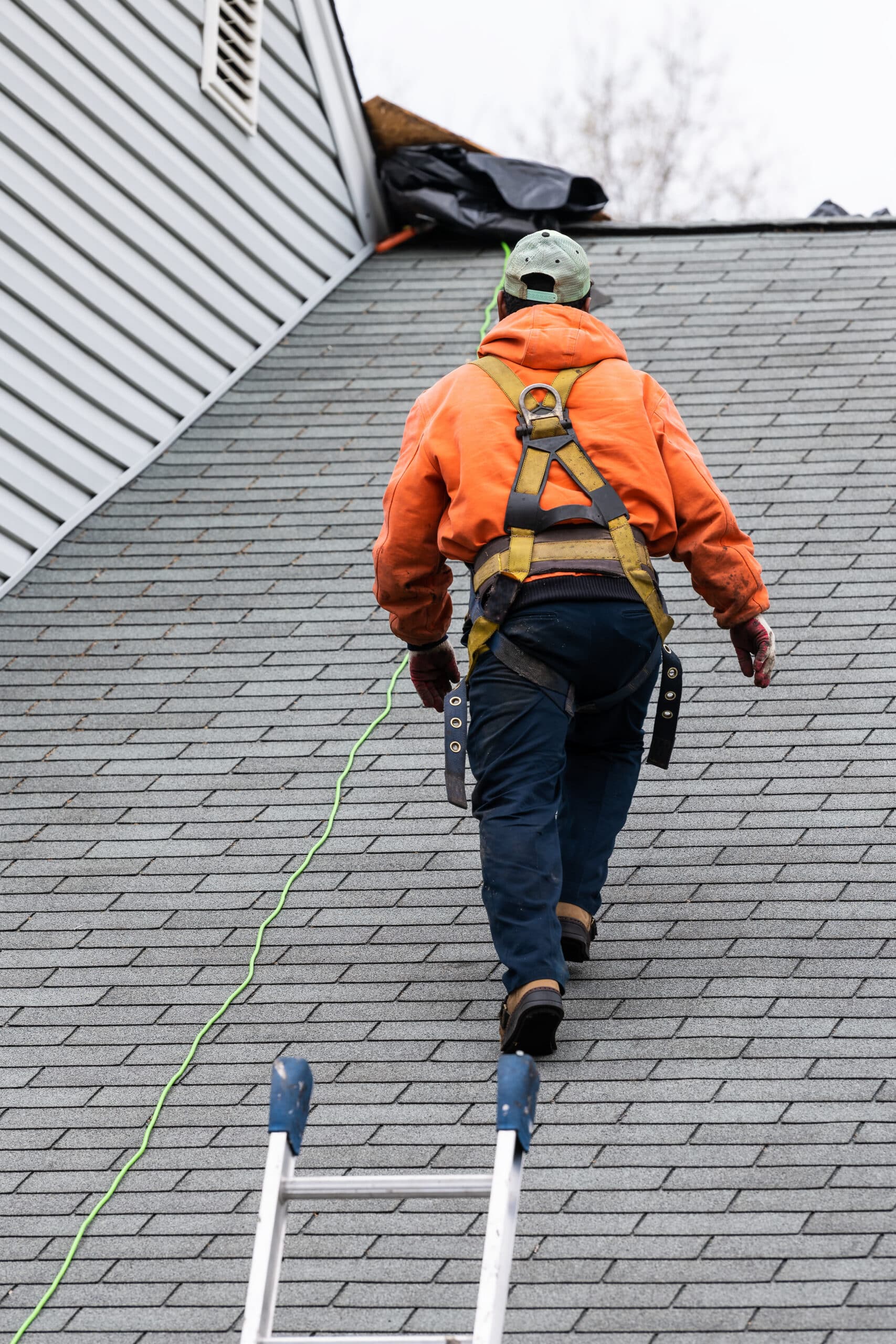 House with gray color Single Family Home and construction man with orange uniform walking on roof shingles and ladder during repair