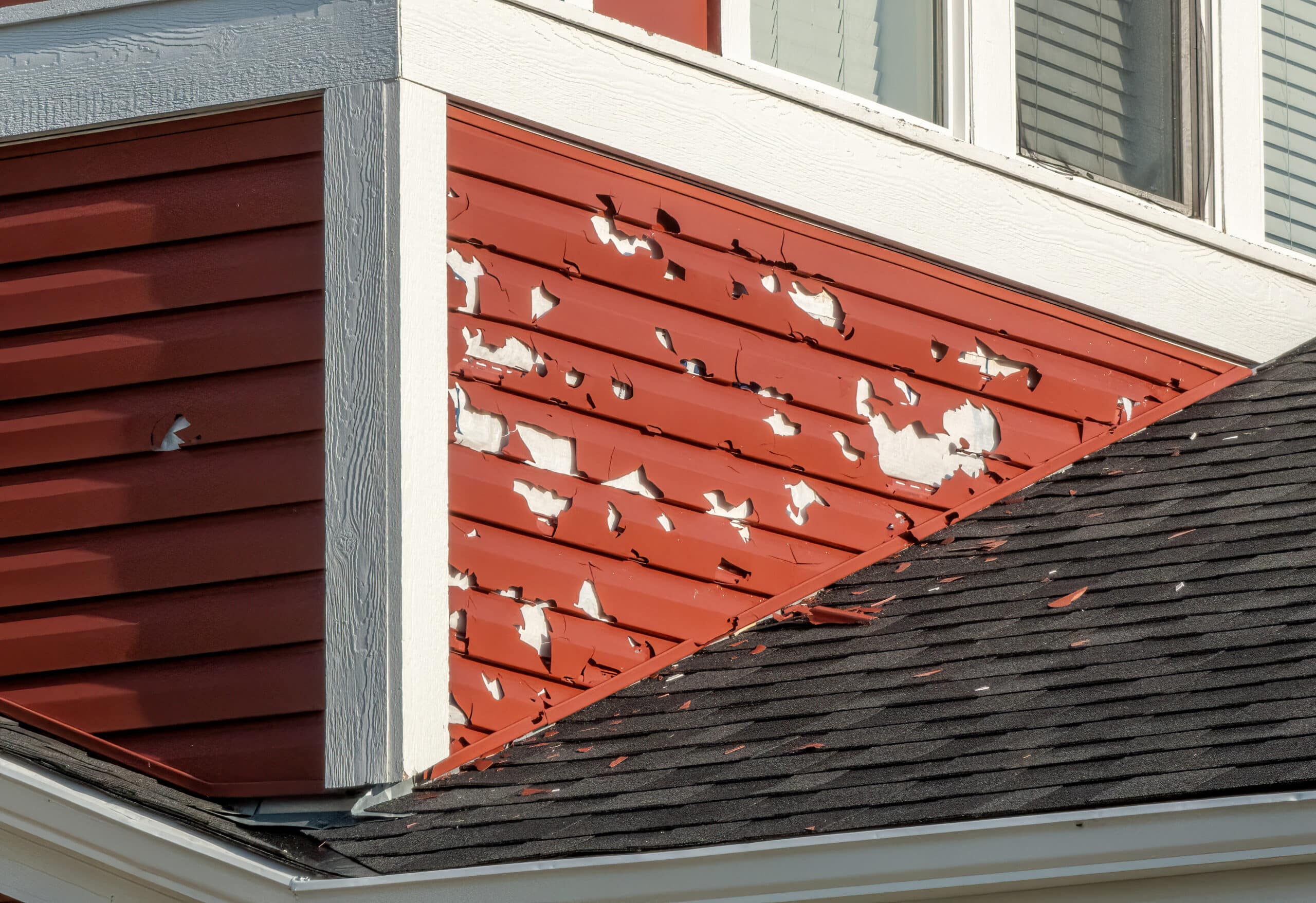 A close-up of a building's exterior shows red siding with extensive hail damage, exposing the underlying white surface.