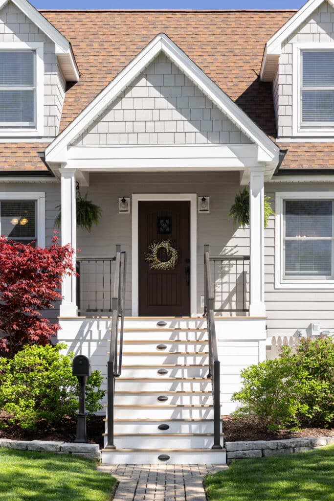 A front door detail with stairs leading to a dark wood front door, covered porch, and a wrought iron railing. Decorative gable roof with white cornice, blue horizontal vinyl