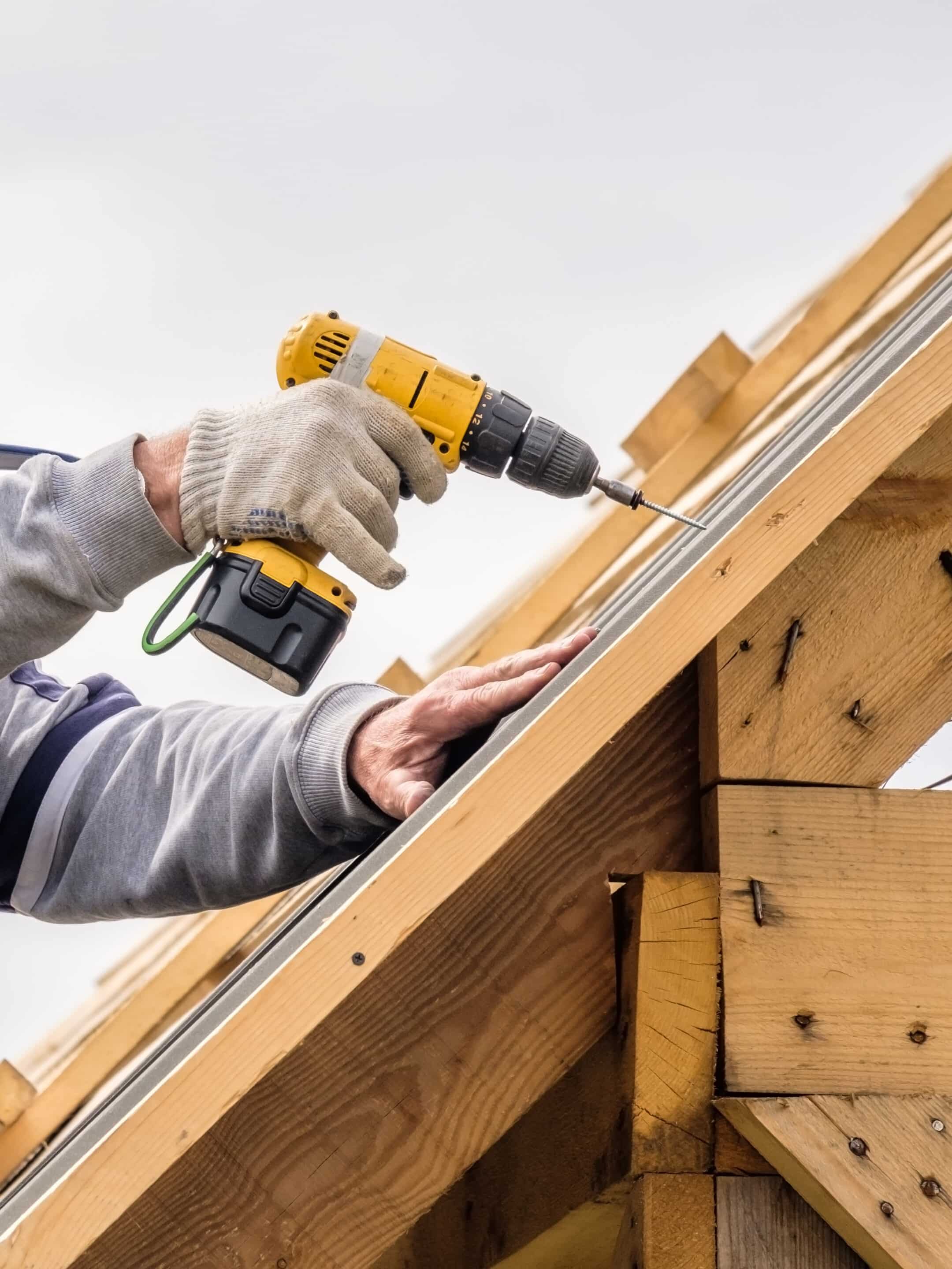 Male hands in gloves with a screwdriver screw the roofing sheet to the roof. Male hands in gloves with a screwdriver screw the roofing sheet to the roof.